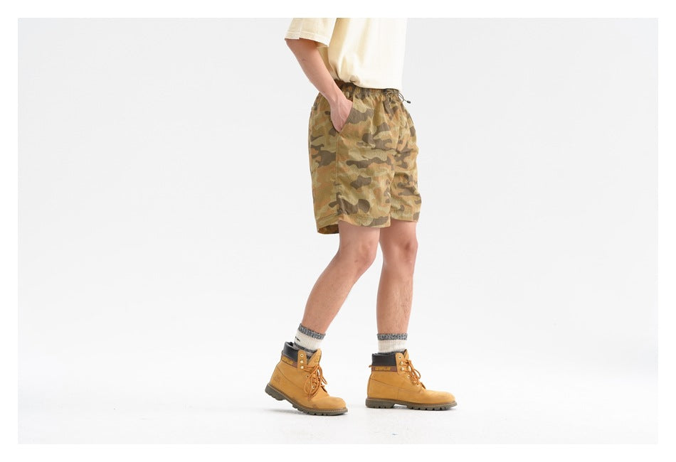 Model wearing olive and brown camouflage drawstring shorts, shown from the side with hands in pockets.
