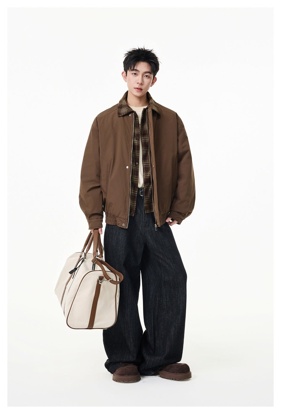 Full-length model wearing a brown jacket with plaid collar and trim, wide dark jeans, and carrying a cream duffel bag.