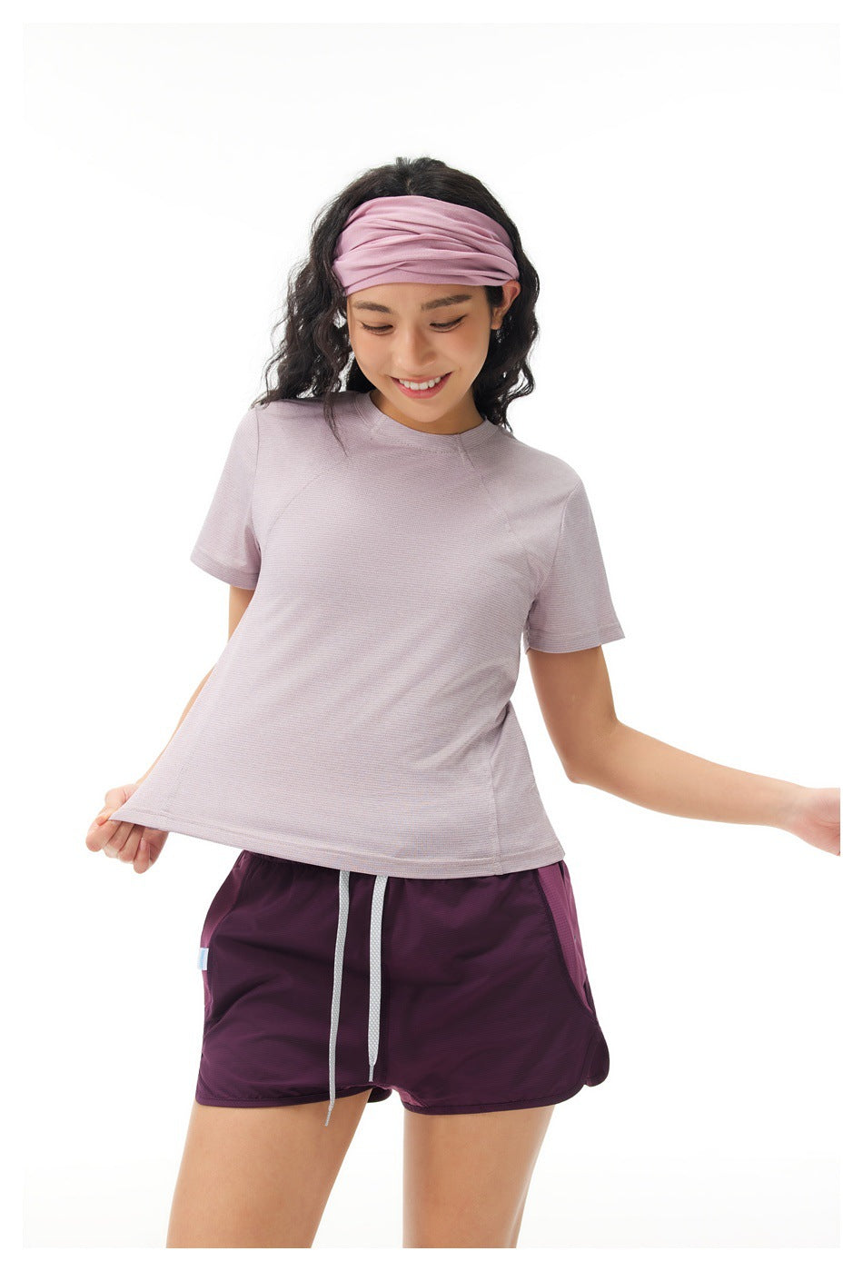 Woman modeling a pale lilac short-sleeve shirt and plum running shorts, holding the shirt hem outward.