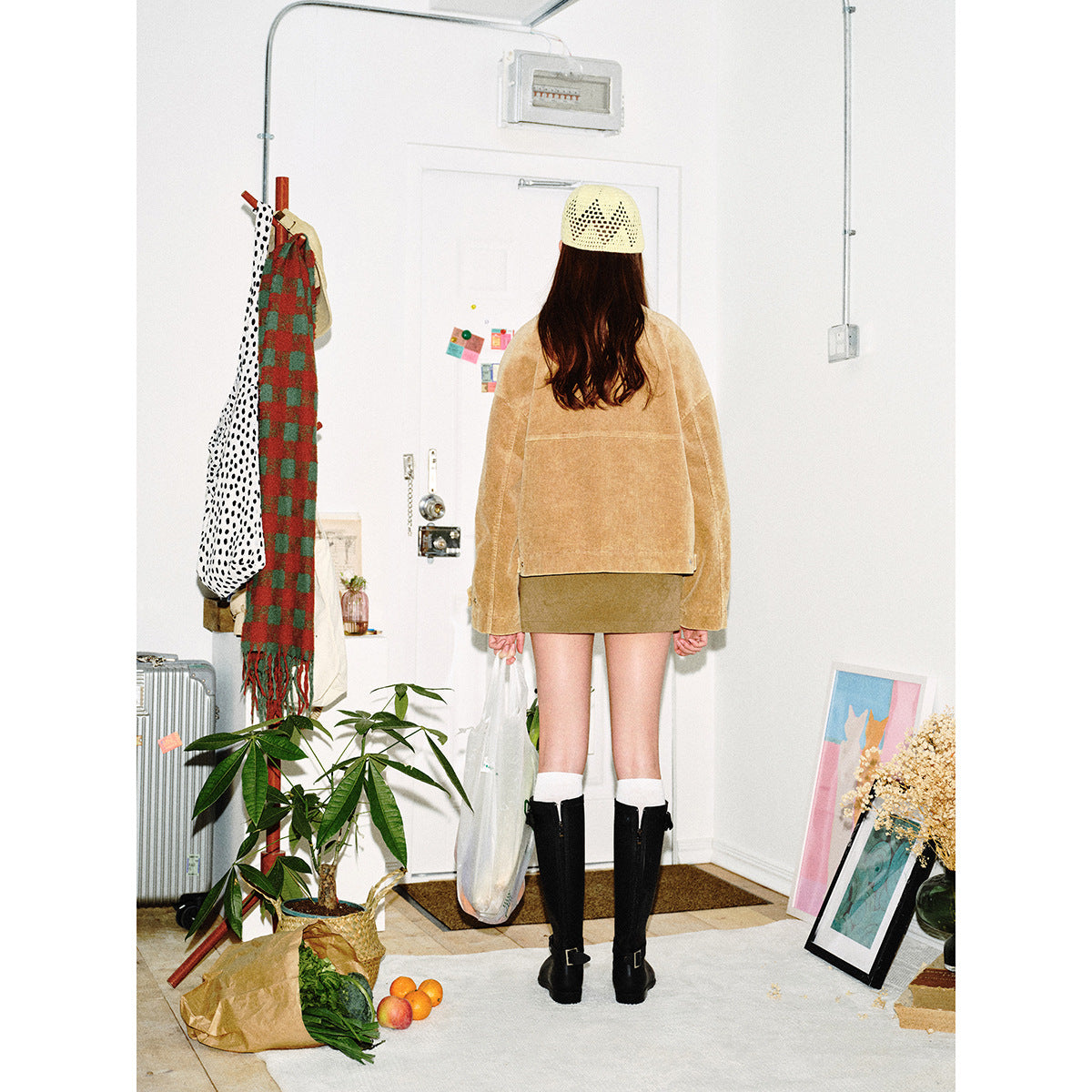Rear view of a model wearing a tan textured jacket, olive mini skirt, cream knit cap, and black boots.