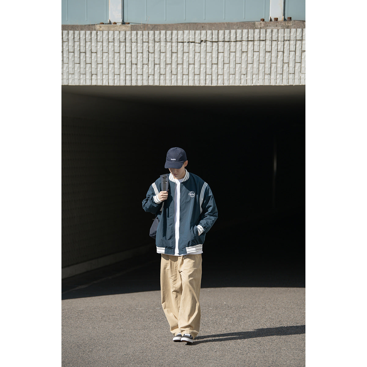 Full-length model wearing a dark navy zip jacket with white striped trim, beige trousers and a navy cap.