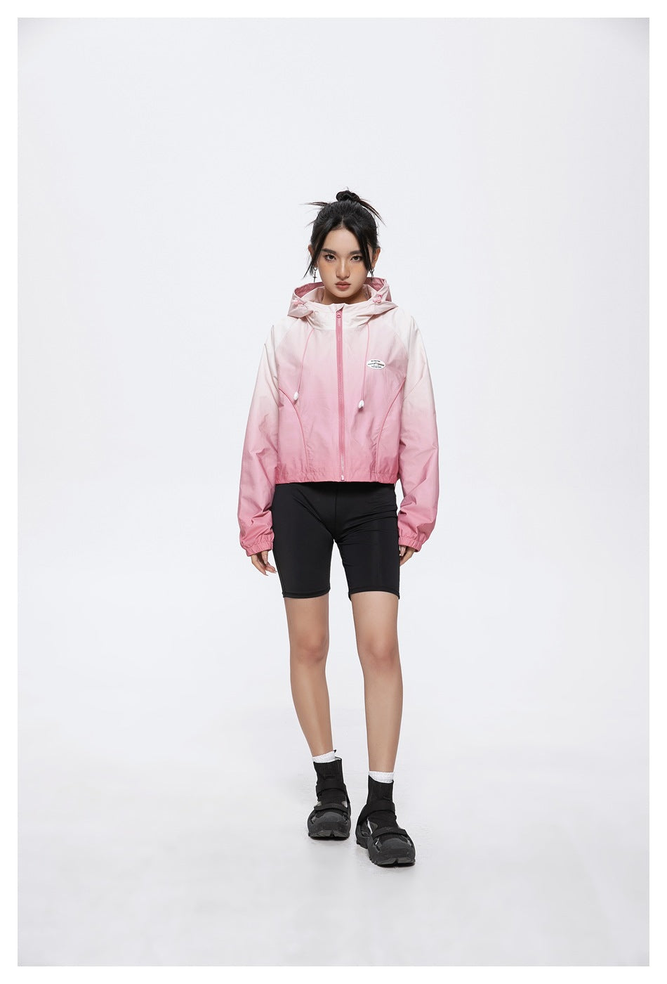 Model wearing a pink gradient hooded zip jacket with black cycling shorts, shown from the front.