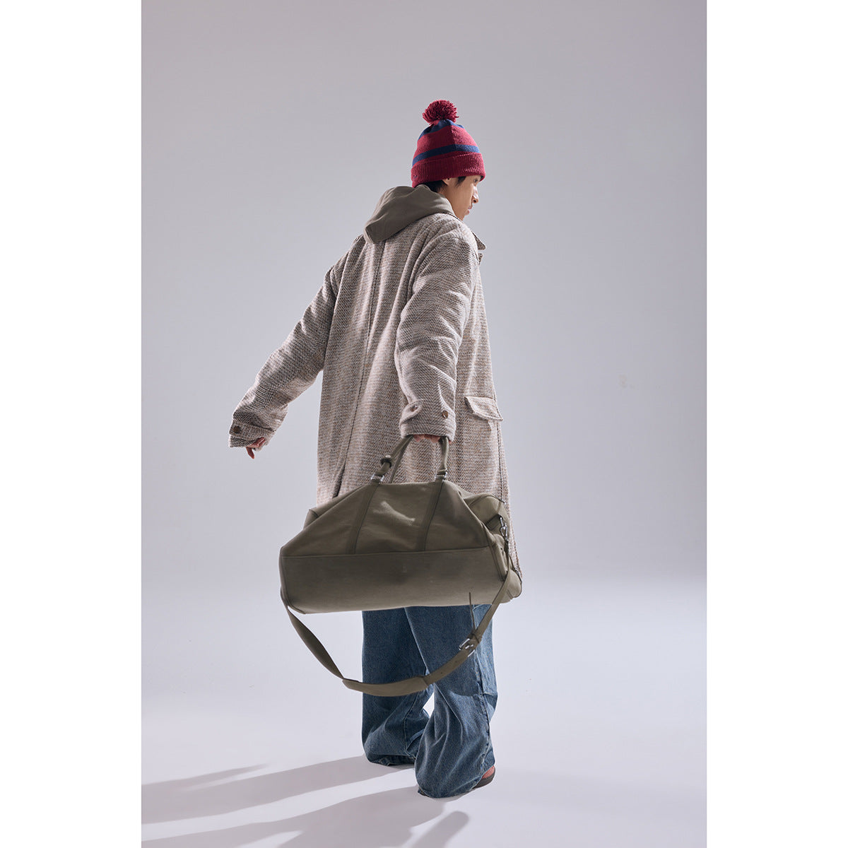 Model shown from behind in a beige patterned long coat, red striped beanie, jeans, and carrying an olive duffel bag.