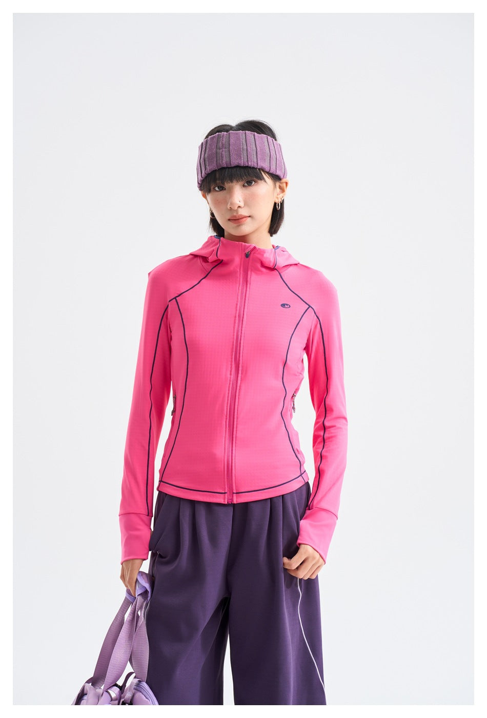 Front view of a model in a bright pink hooded zip jacket with dark piping and loose purple pants.