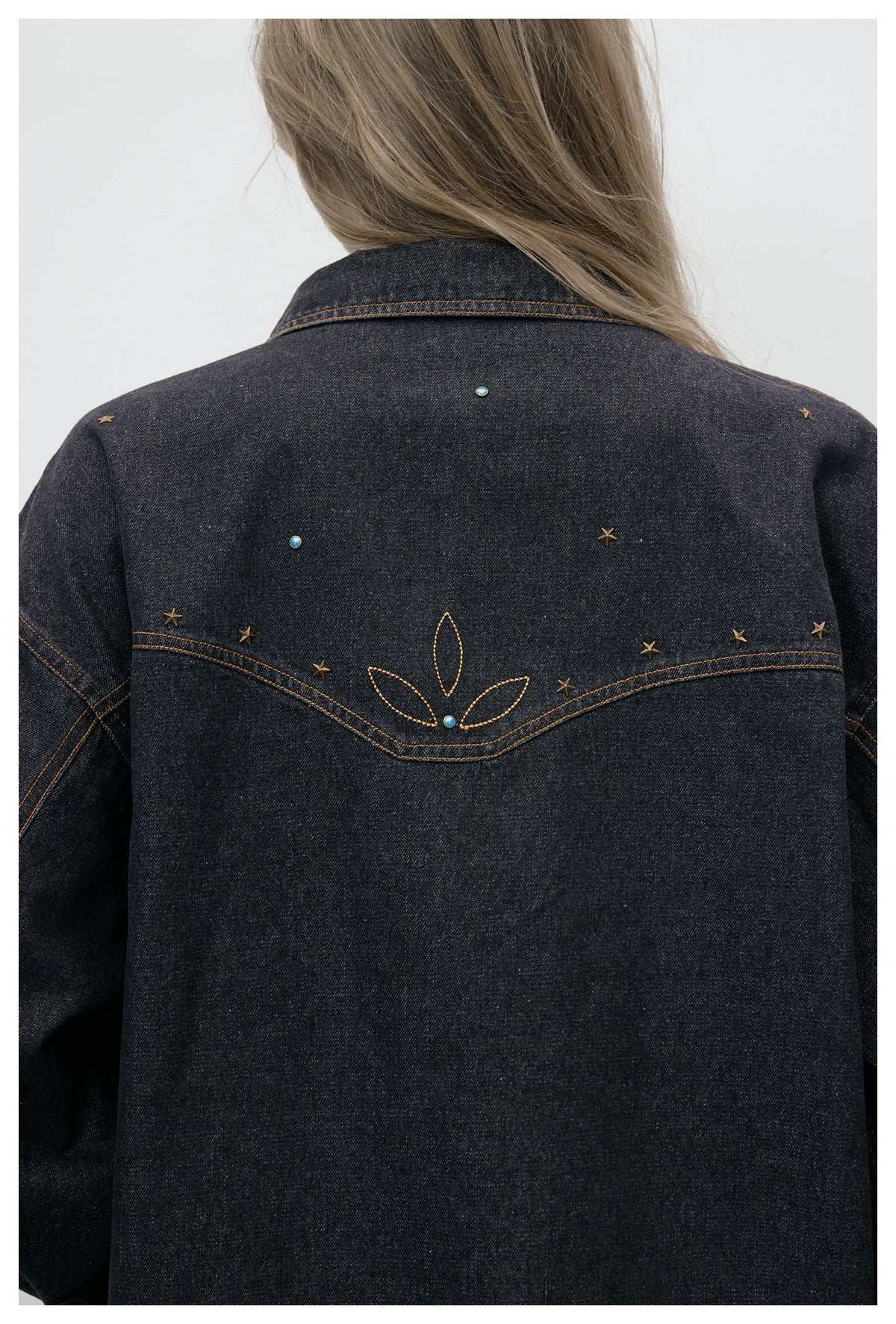 Close rear view of a charcoal denim jacket with bronze star studs, turquoise accents, and curved floral stitching.