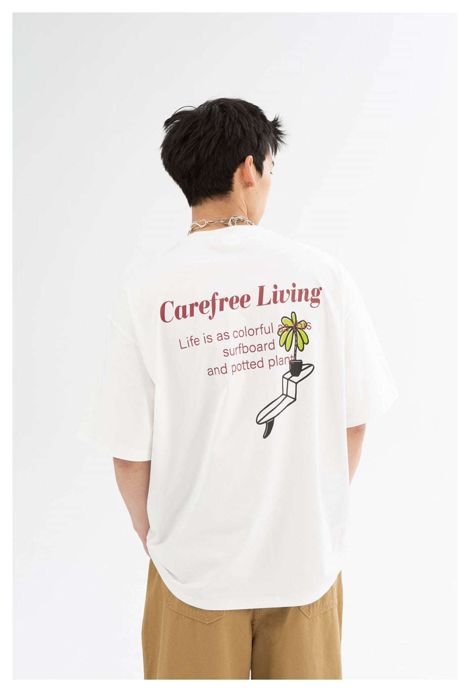 Back view of a model wearing a white T-shirt with colorful surfboard and plant artwork and tan trousers.