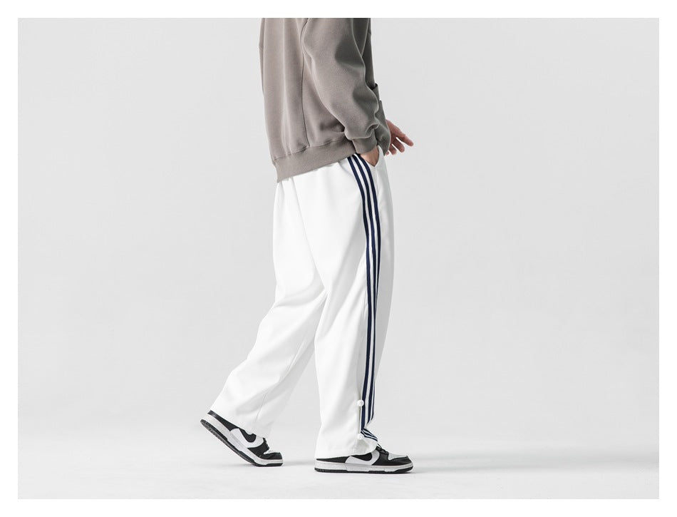 Rear side view of a model wearing loose white track pants with three navy side stripes and snap hems.