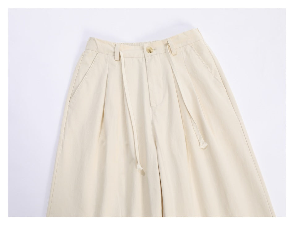Close view of ivory trousers showing the front waistband, button, belt loops, pleats, angled pockets and drawstrings.