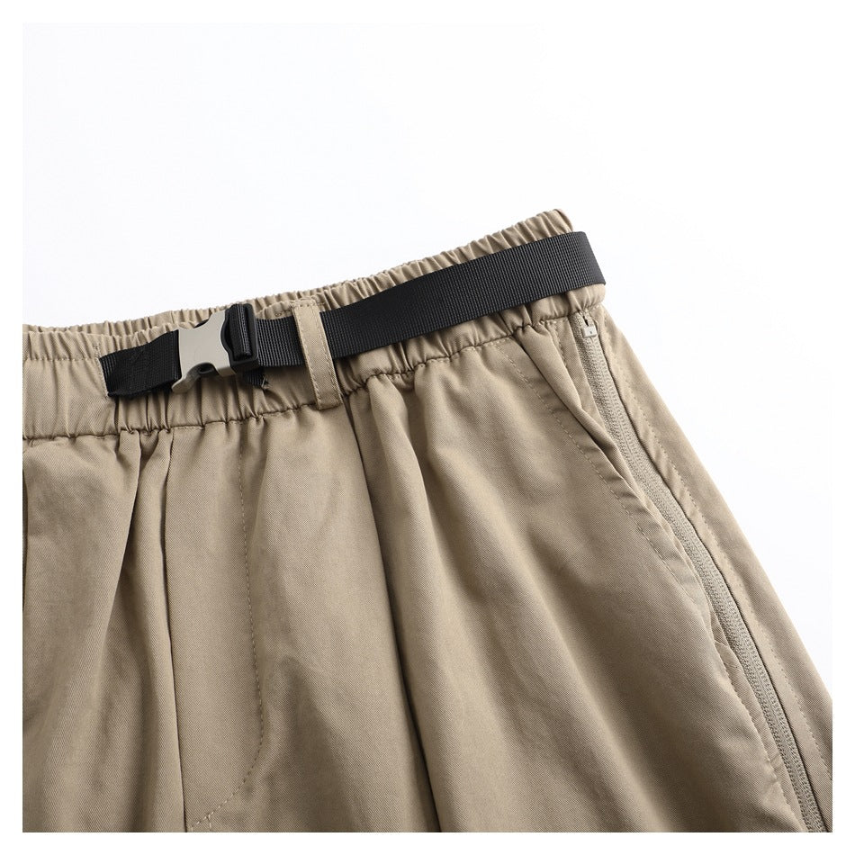 Angled close-up of taupe trousers showing the elastic waistband, black adjustable belt and zip pocket.