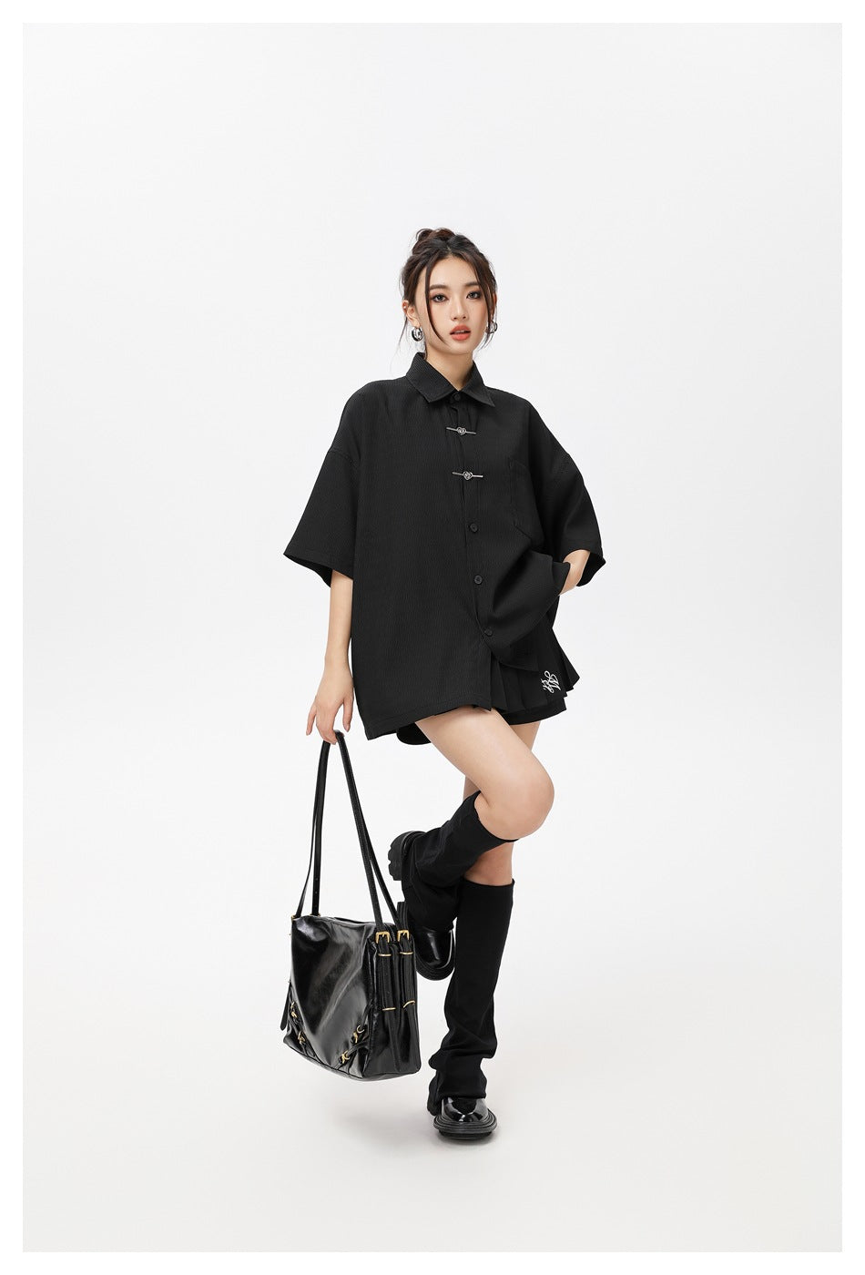 Model wearing a black oversized short-sleeve shirt with metal toggles, styled with a black handbag and tall socks.