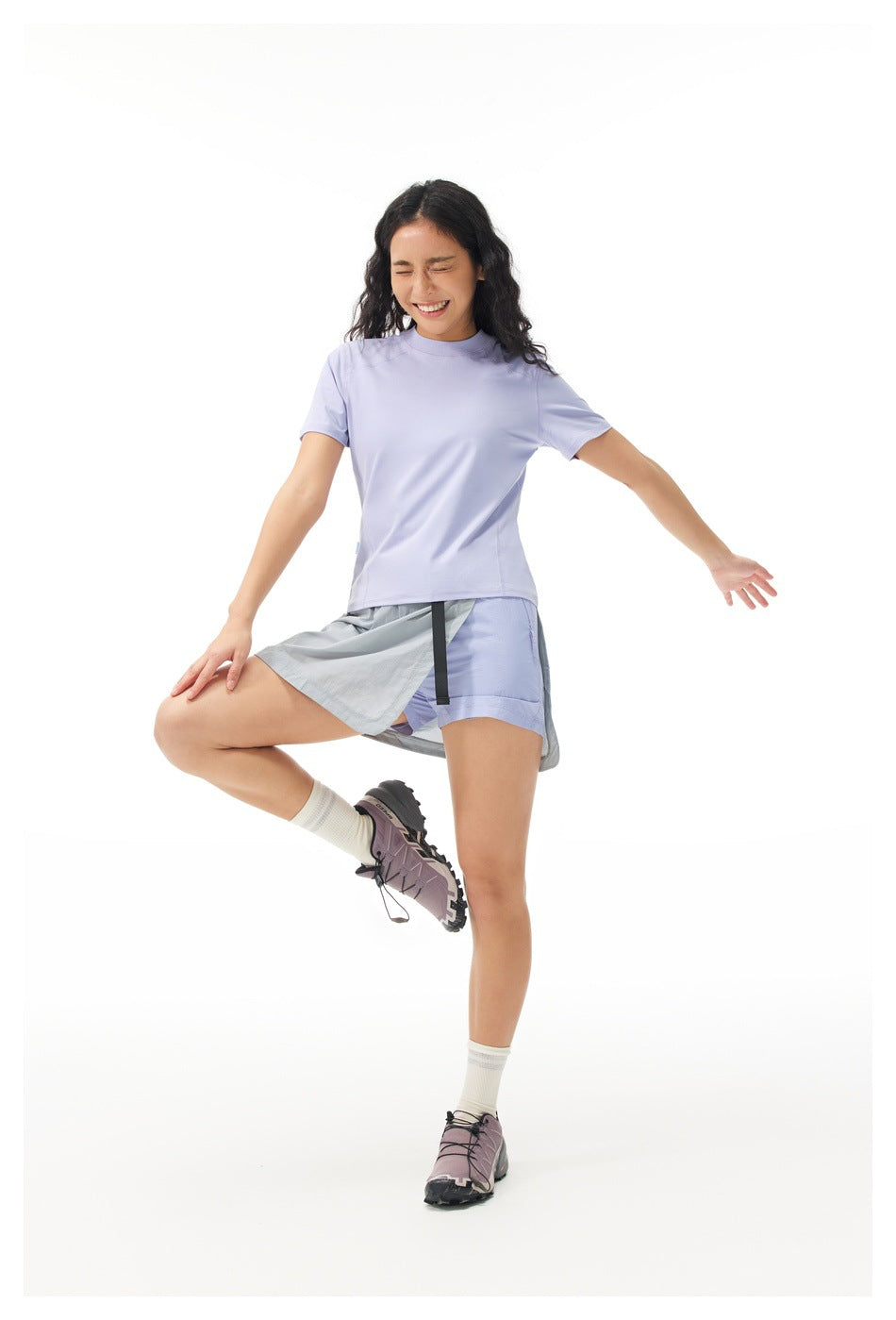 Model wearing a lavender T-shirt and gray wrap-style tennis skirt over matching shorts, lifting one leg.