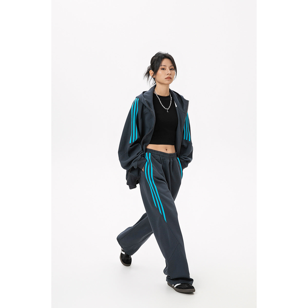 Model wears an open charcoal hoodie and loose matching pants with bright cyan stripes and a black crop top.