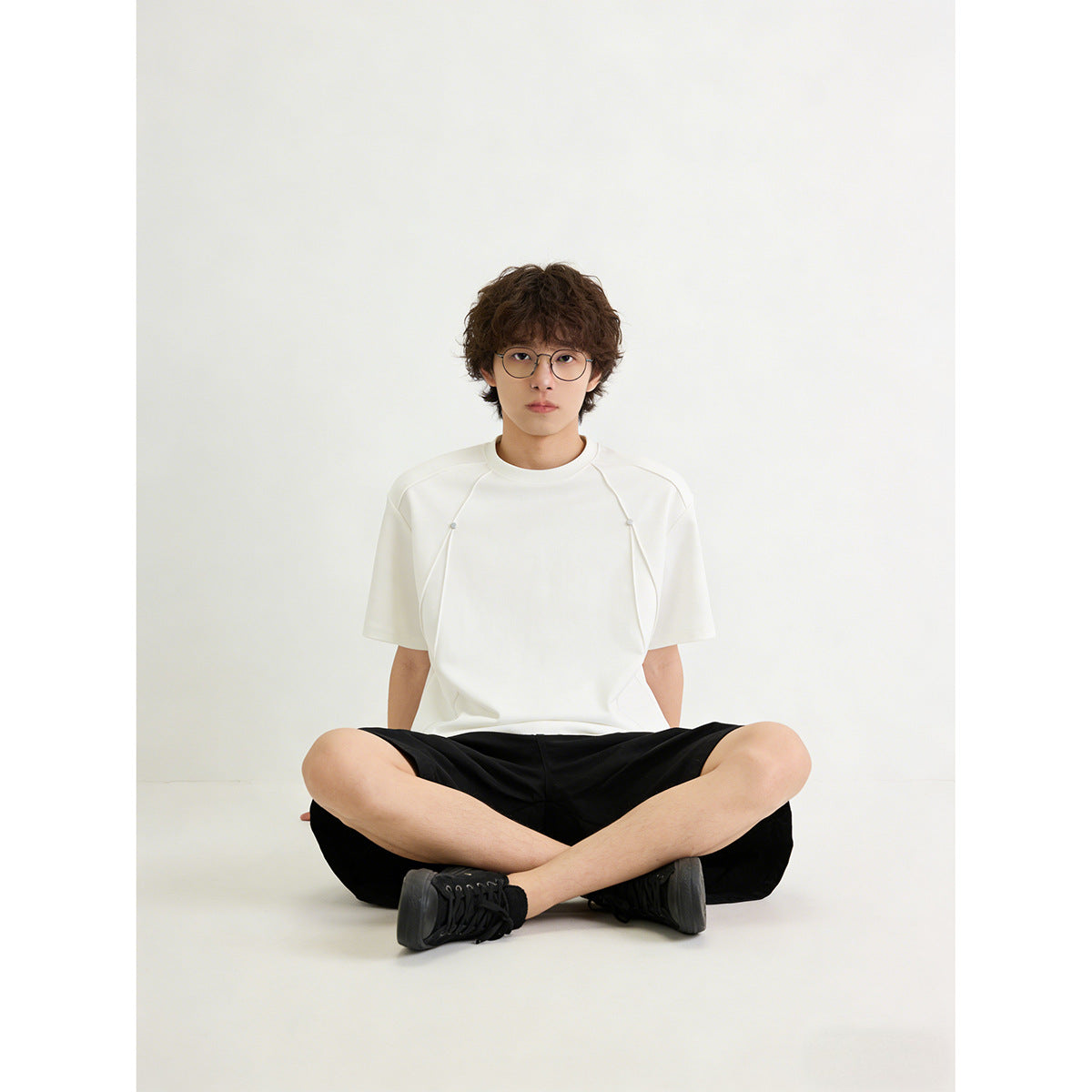 Model seated cross-legged wearing an oversized white short-sleeve tee with curved front panels and small snaps.