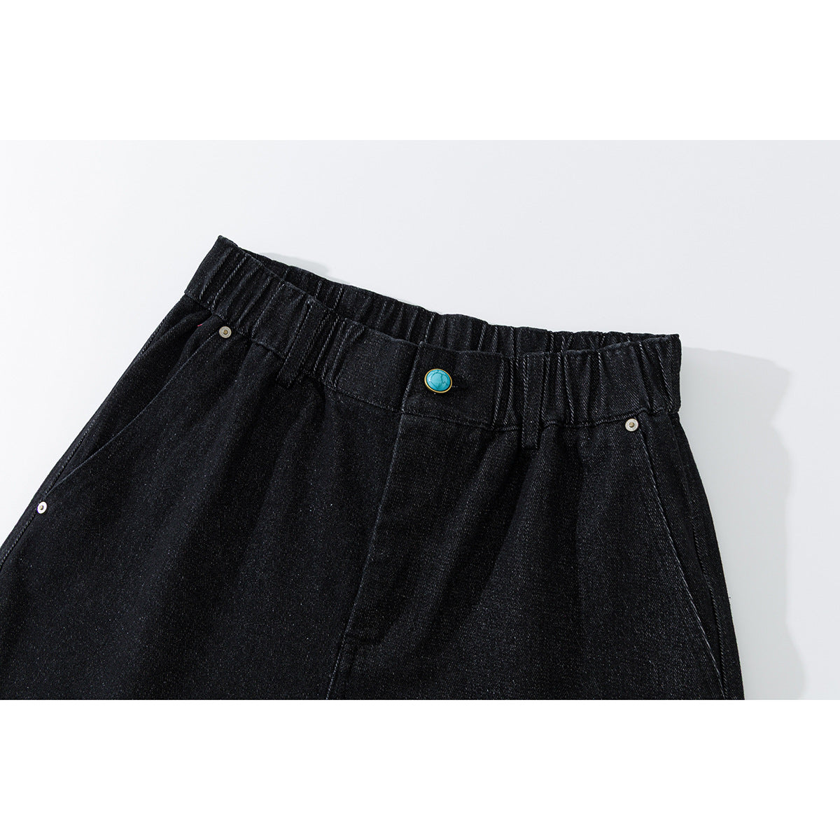 Close-up of black denim shorts with an elastic waistband, turquoise button, belt loops and riveted pockets.