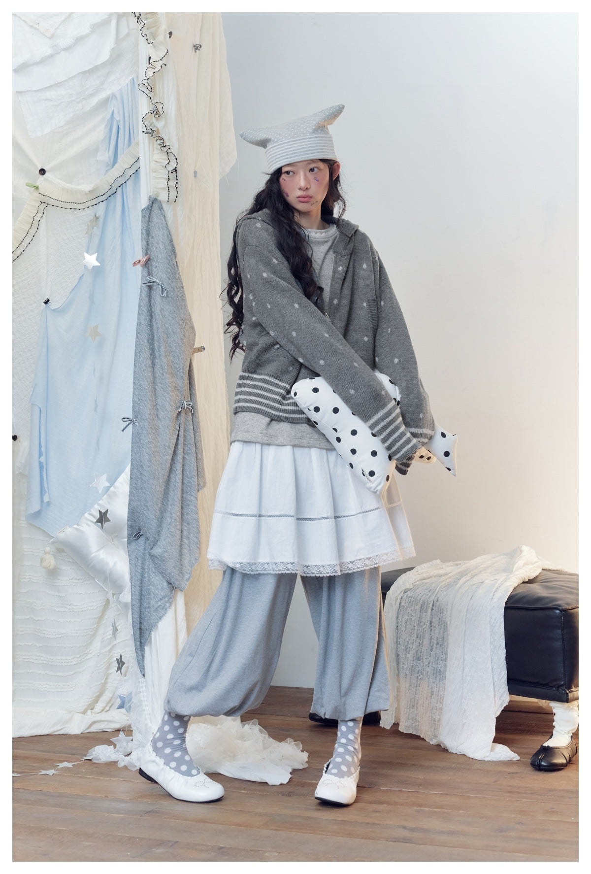 Model turned sideways in gray polka-dot hooded knit jacket, holding a dotted pillow, with white skirt and gray pants