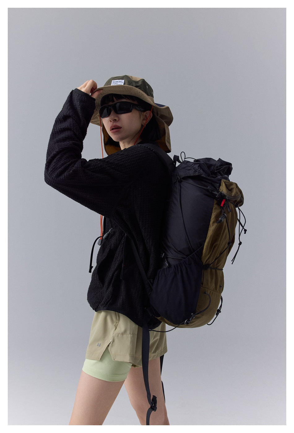 Model wearing a black textured zip jacket with khaki shorts and a large hiking backpack.