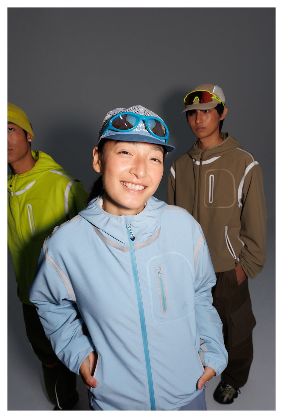 A model in a pale blue hooded jacket stands in front of models wearing neon yellow-green and tan versions.
