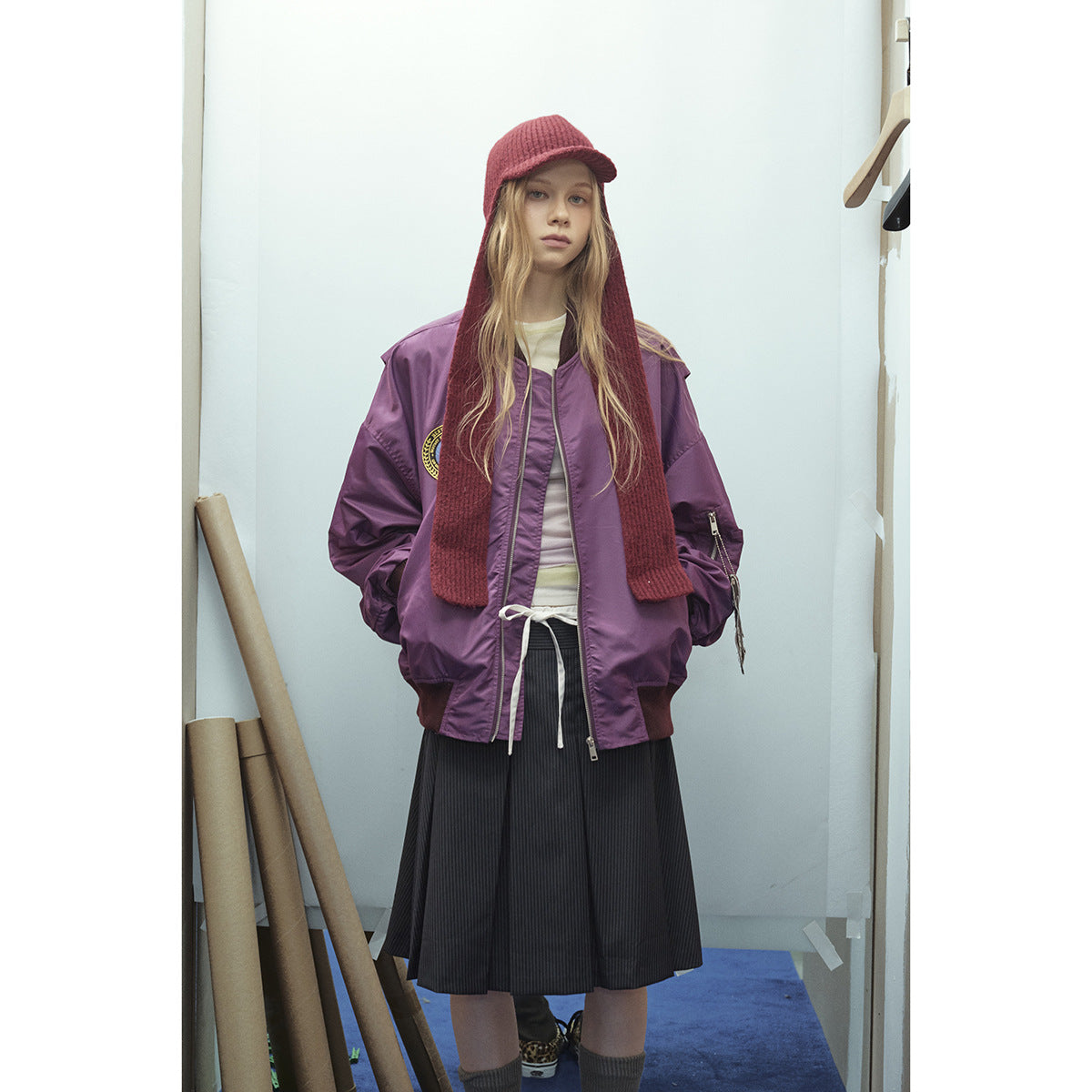 Model wearing a purple zip-front bomber jacket with a chest patch, burgundy knit hood scarf, and black pleated skirt.