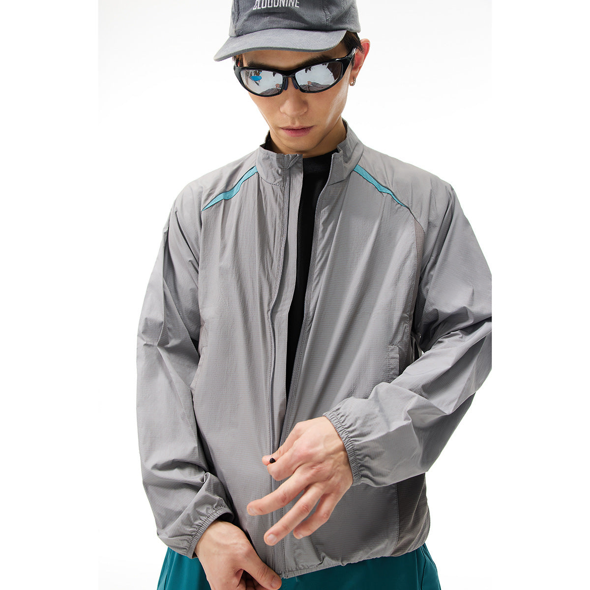 Close view of a man wearing a gray zip jacket with turquoise shoulder accents; he wears a cap and sunglasses.