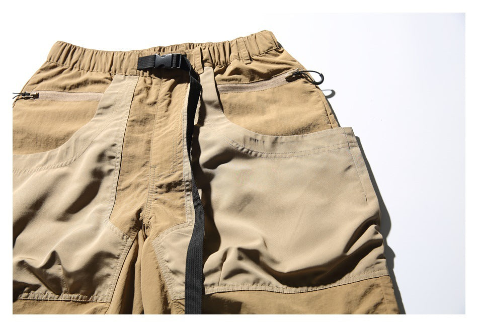 Close-up of the front of tan utility shorts with beige patch pockets, zipper pockets, and a black buckle belt.