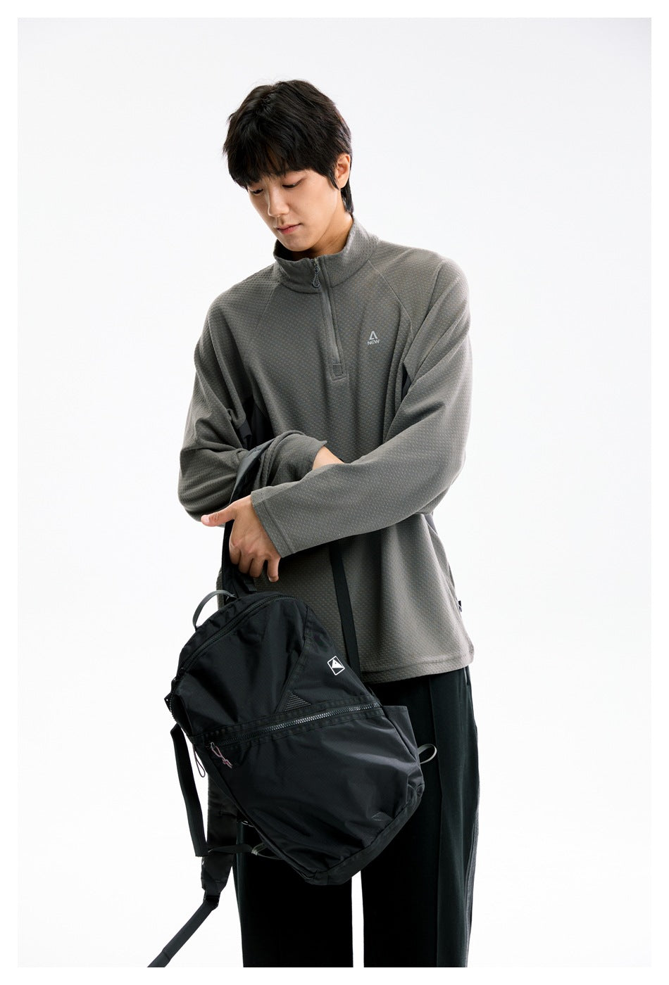 Model wearing a textured gray quarter-zip pullover and holding a black backpack against a white studio background.