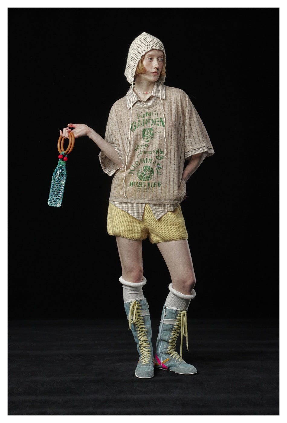 Model wearing a beige green-graphic tee over a checked shirt, yellow shorts, a cream crochet bonnet, and tall blue boots.
