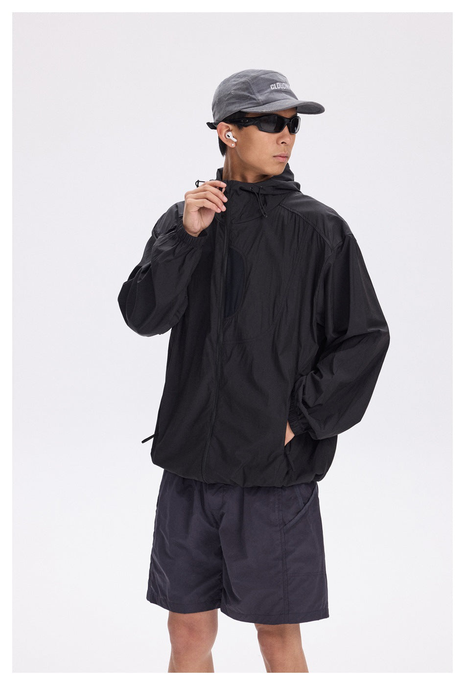 Model wearing a black hooded jacket and dark shorts, shown with a gray cap and sunglasses.