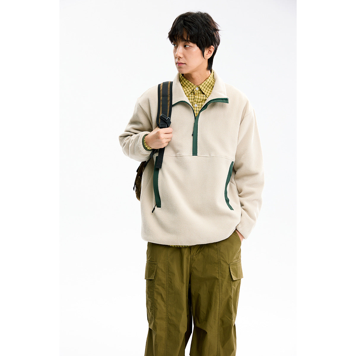 Model wearing a cream pullover with dark green trim over a yellow checked shirt, with olive cargo pants and a backpack.