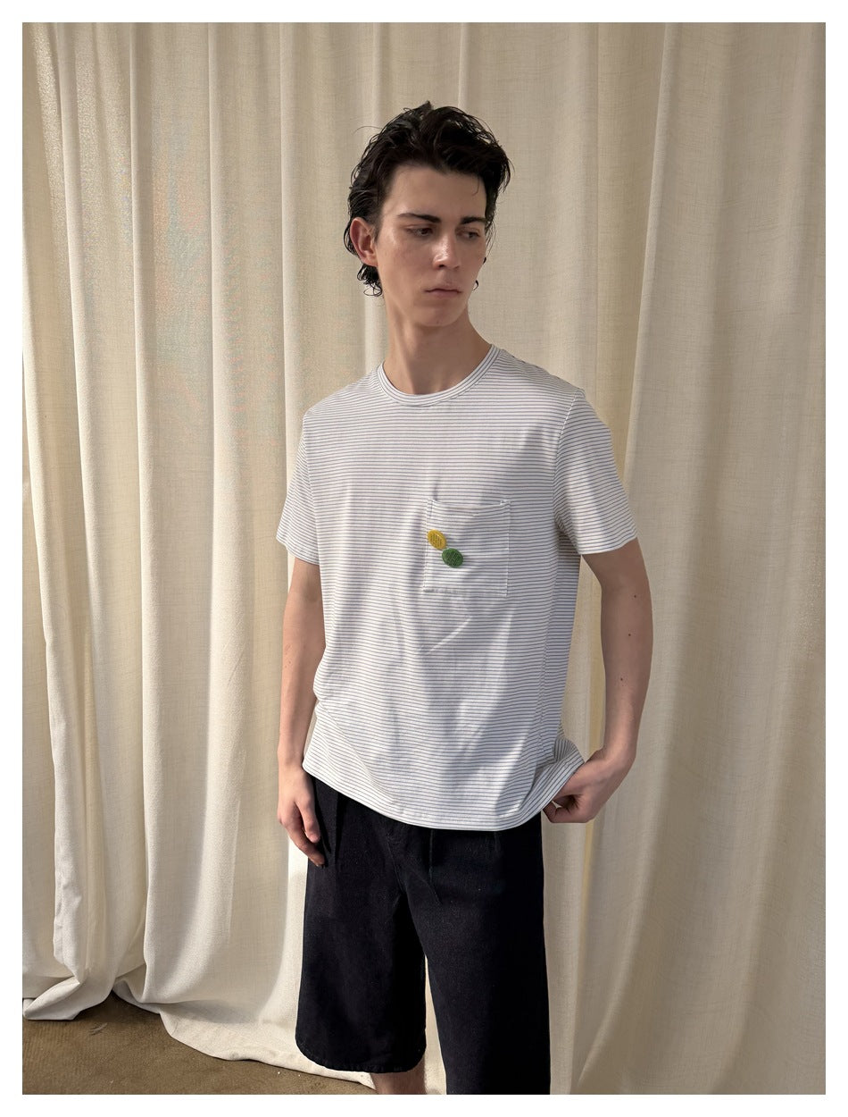 Model wearing a white T-shirt with fine blue stripes, a chest pocket with two small colorful accents, and black shorts.