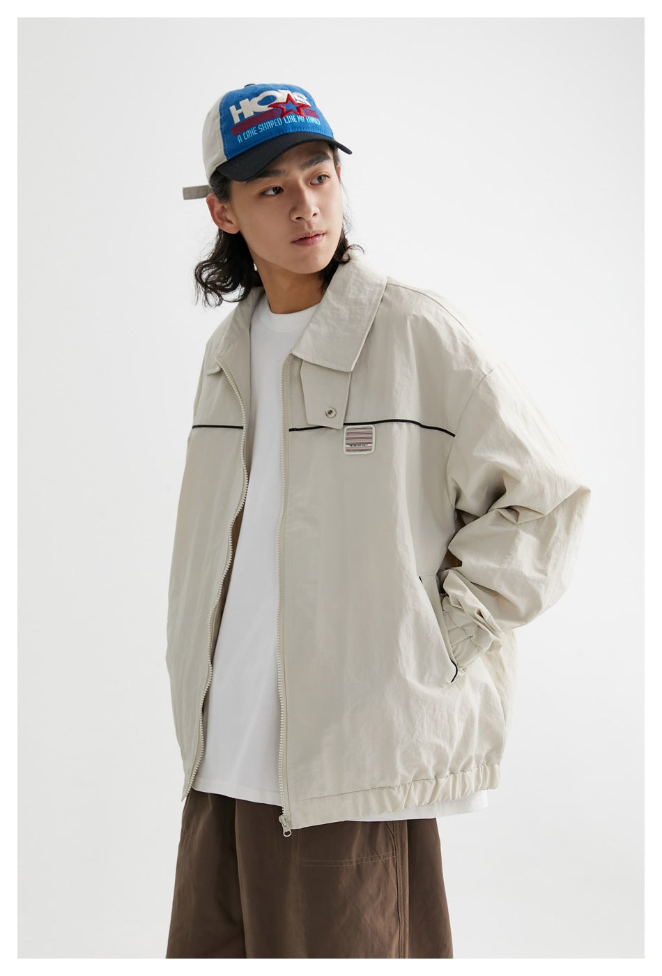 Model wearing a beige zip jacket with black piping and chest patch over a white tee, with a hand in the pocket, close-up