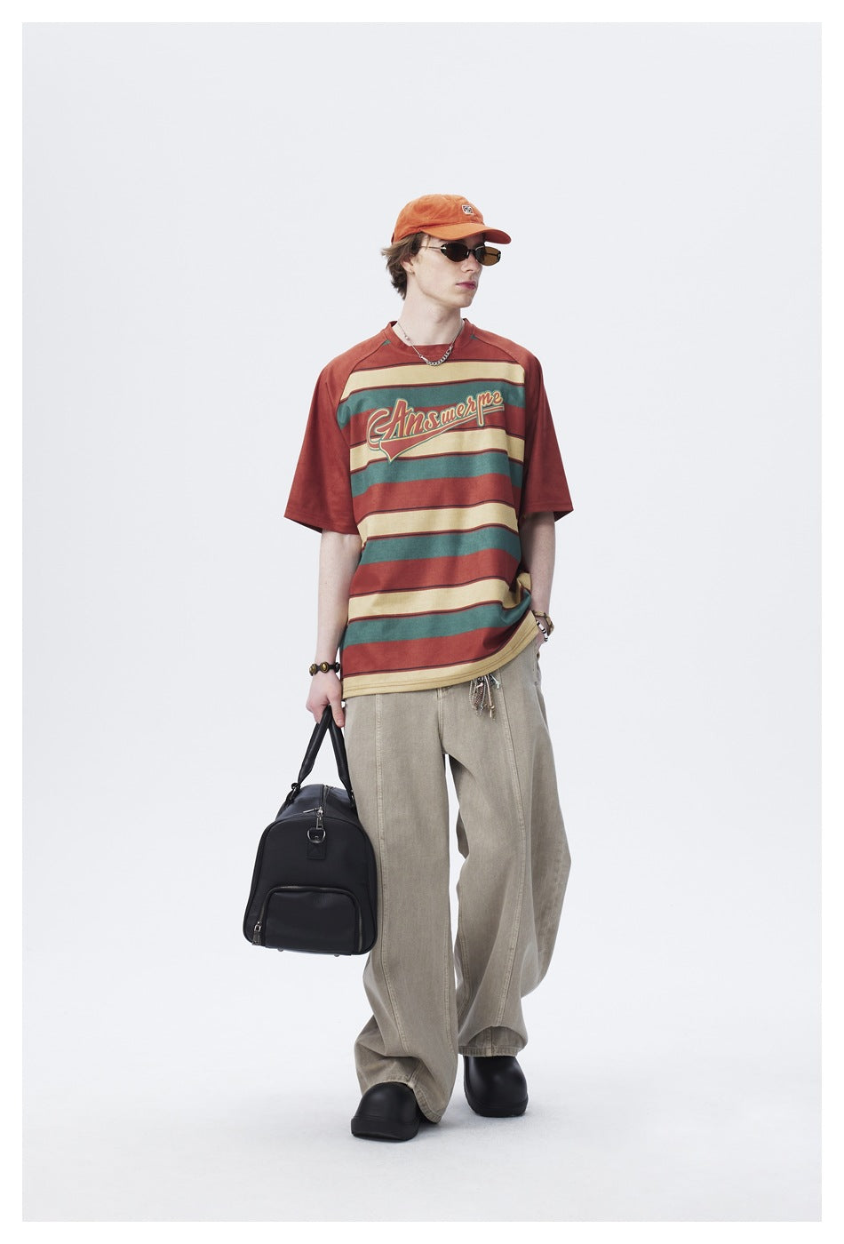 Full-length model wearing a rust, teal and cream striped graphic T-shirt with loose beige trousers.