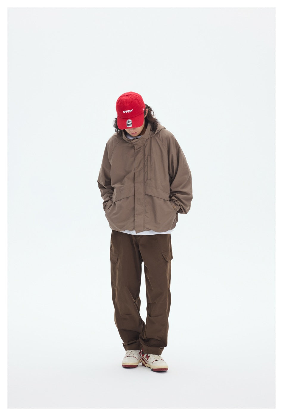 Model wears a taupe hooded jacket with a covered front closure and dark cargo pants, head tilted down.