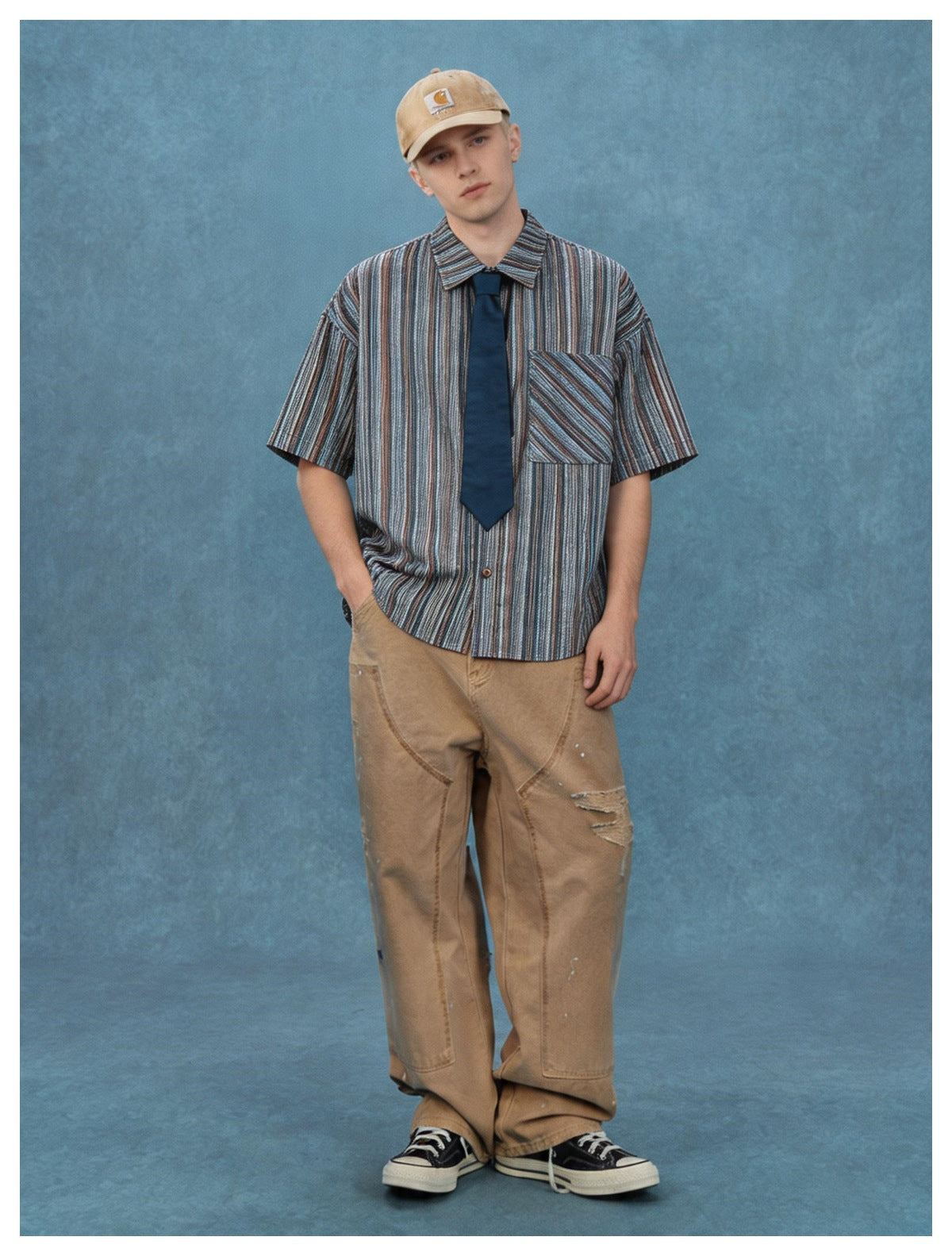 Man wearing navy striped short-sleeve shirt with tie, cap, tan wide-leg pants and sneakers