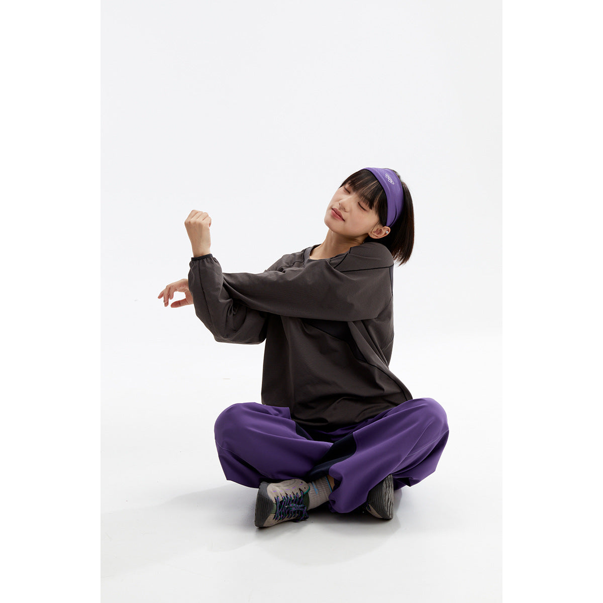 Woman seated in a studio wearing a charcoal long-sleeve top, purple pants, and a matching purple headband.