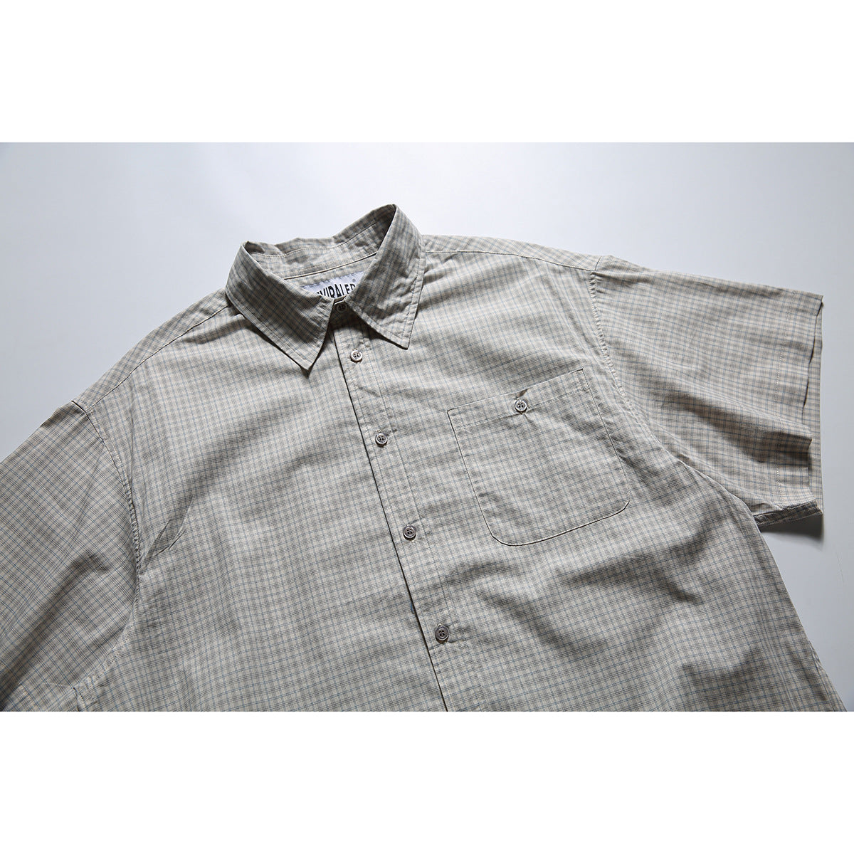 Flat-lay front view of a light gray short-sleeve shirt with a fine blue and brown plaid pattern and buttoned chest pocket.