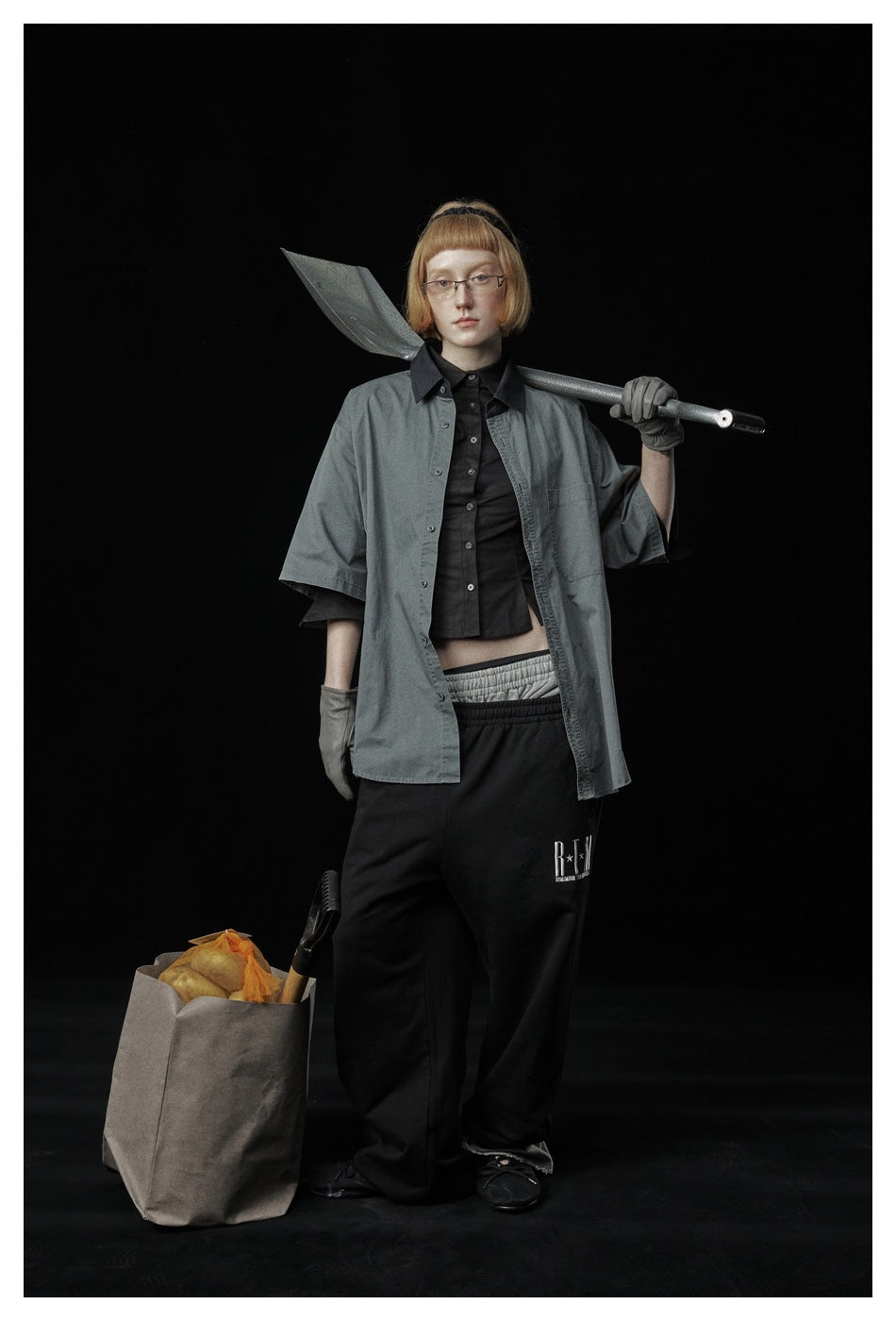 Person wearing the gray short-sleeve shirt open over a black shirt, with a shovel on the shoulder