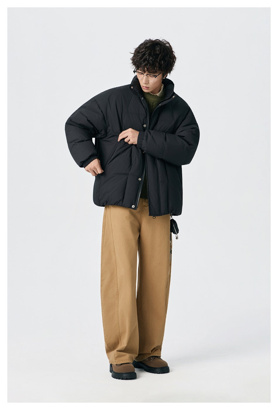 Model adjusting the front of a black puffer jacket with tan trousers.