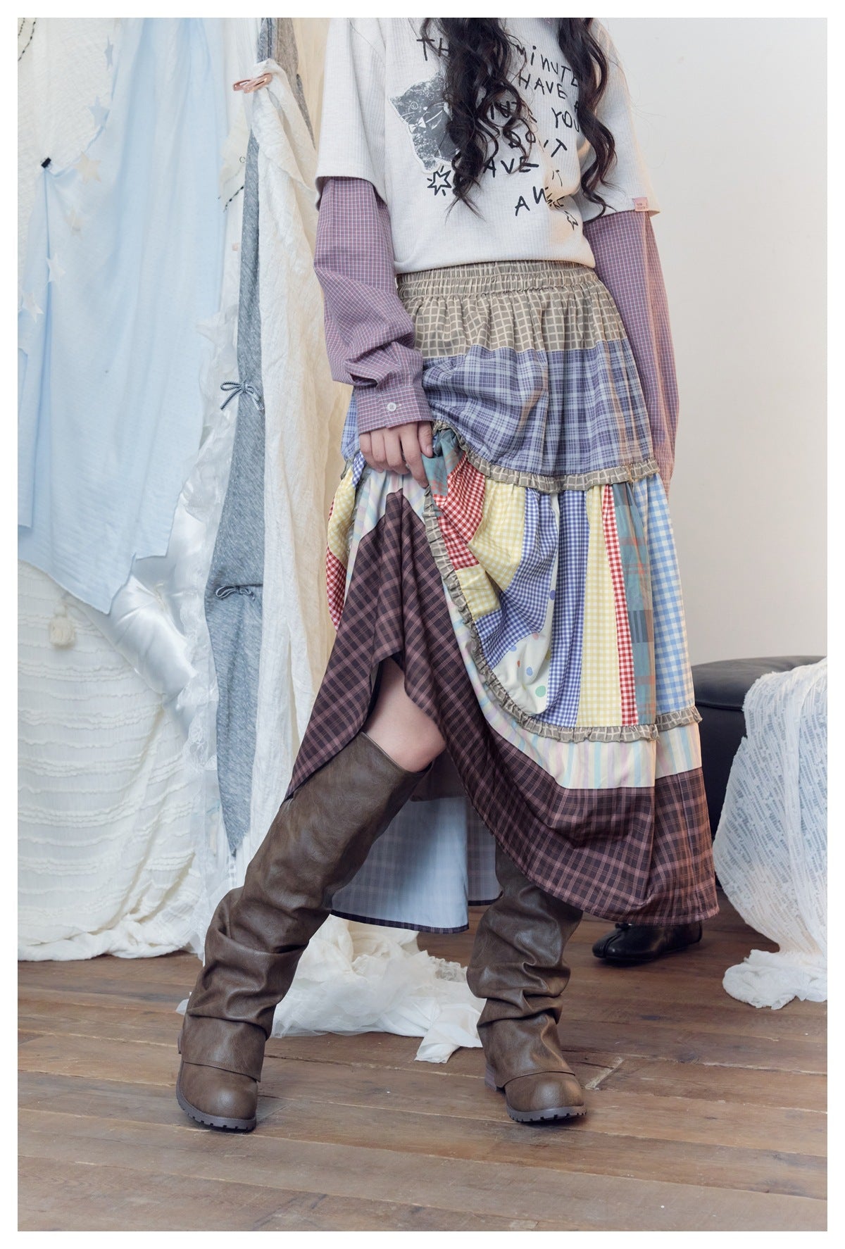 Person lifting the hem of the tiered patchwork skirt to show the plaid layers, worn with brown boots