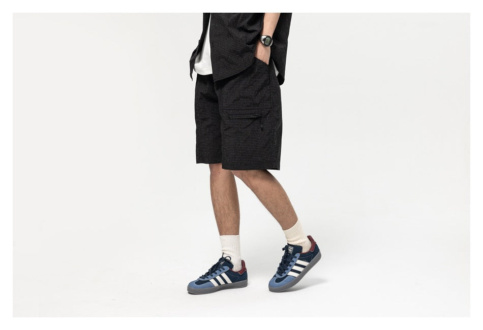 Side view of a model in black grid-pattern shorts with a zip thigh pocket, hand in pocket, wearing a watch and sneakers