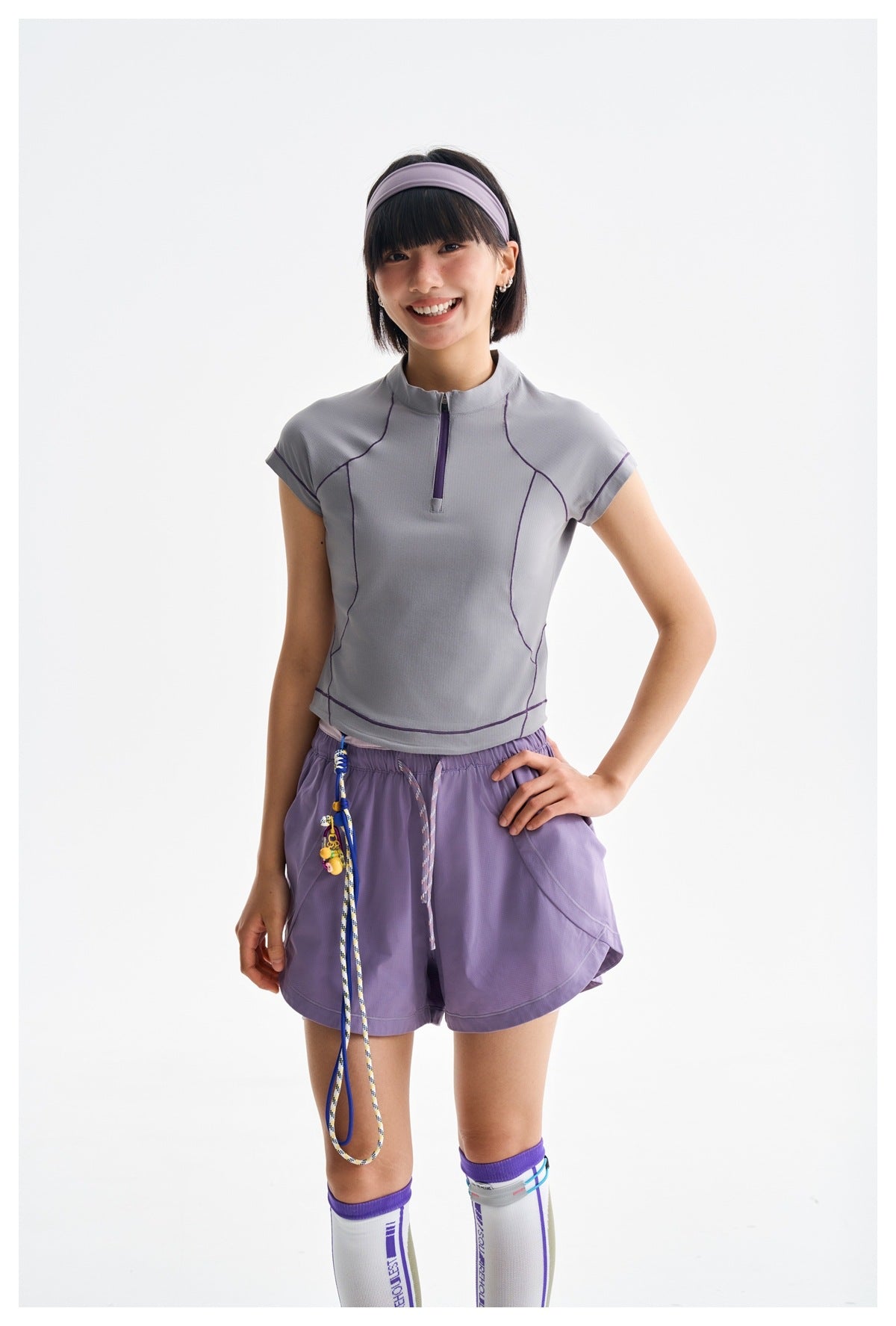 Model wearing a light gray-lilac zip-neck top with purple seam accents and lilac shorts.