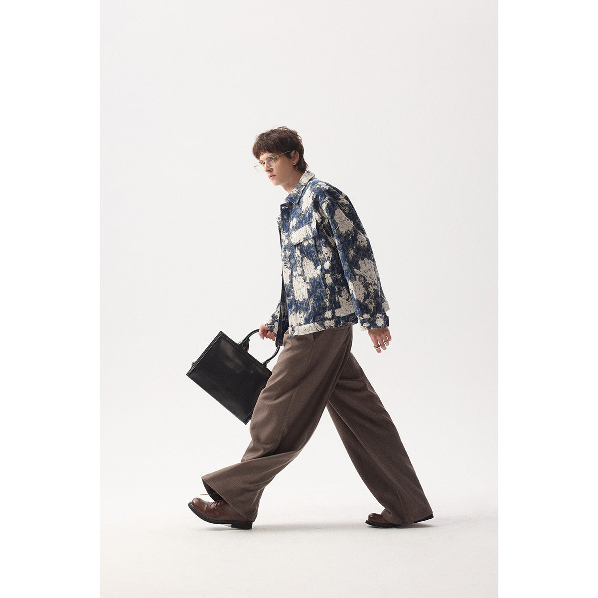 Model walking in a blue and cream floral jacket, wide brown trousers, and brown shoes, carrying a black bag.