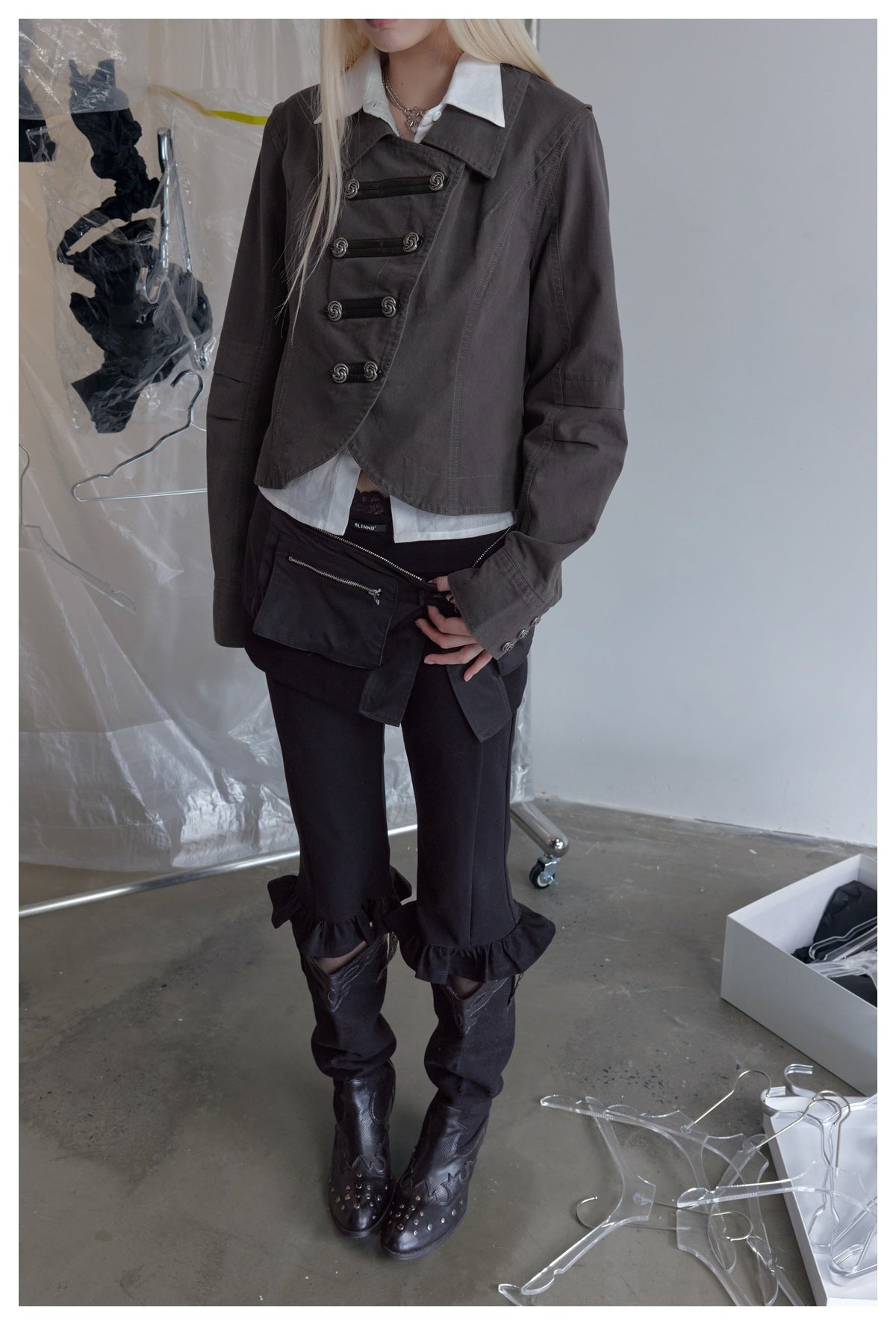 Model wearing a charcoal cropped jacket with an asymmetrical front and ornate toggles, over a white collared shirt.