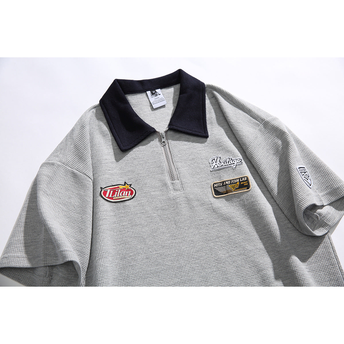 Flat-lay front of a light gray textured short-sleeve polo with a dark collar, quarter zipper, and embroidered chest and