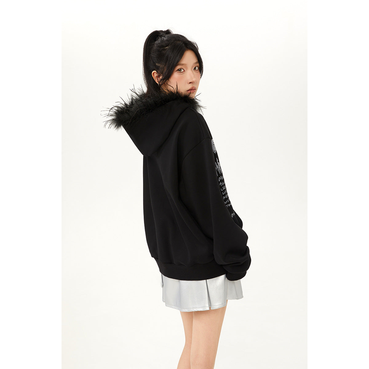 Model showing the back and side of a black oversized hoodie with fur-trimmed hood, worn with a silver skirt.