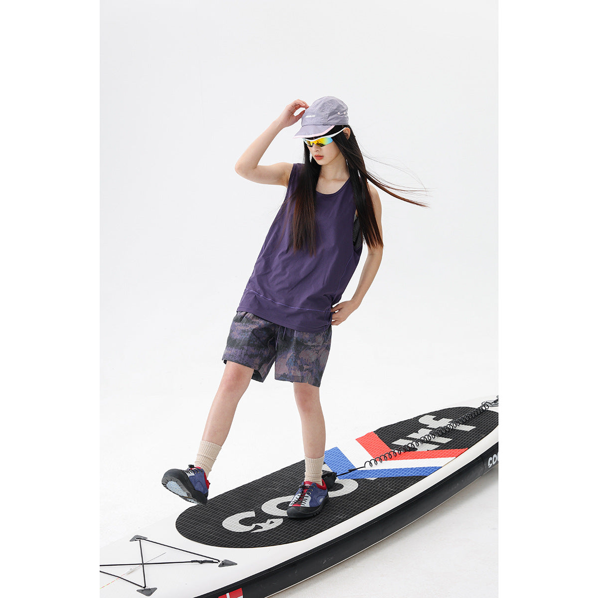 Model in a purple sleeveless tank and patterned dark shorts, standing on a paddleboard.