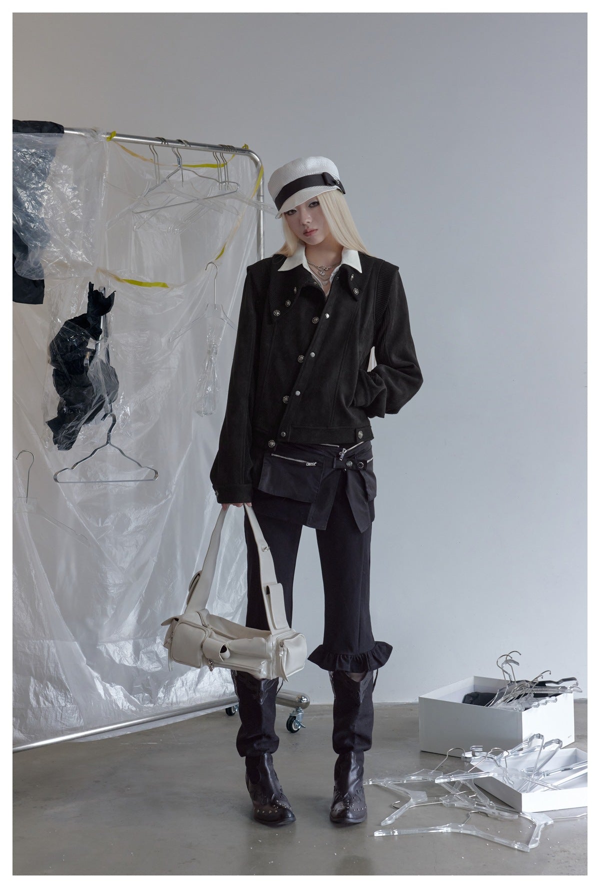 Model wearing the black cropped jacket with a white cap, white collared shirt, black pants and boots, holding a white bag