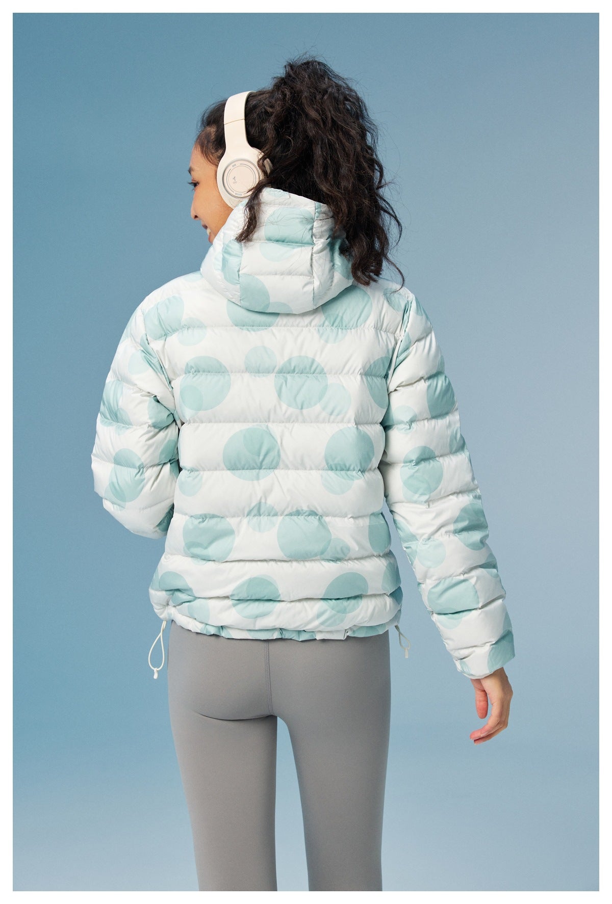 Rear view of a model wearing a white hooded puffer jacket with large pale aqua circles and gray leggings.