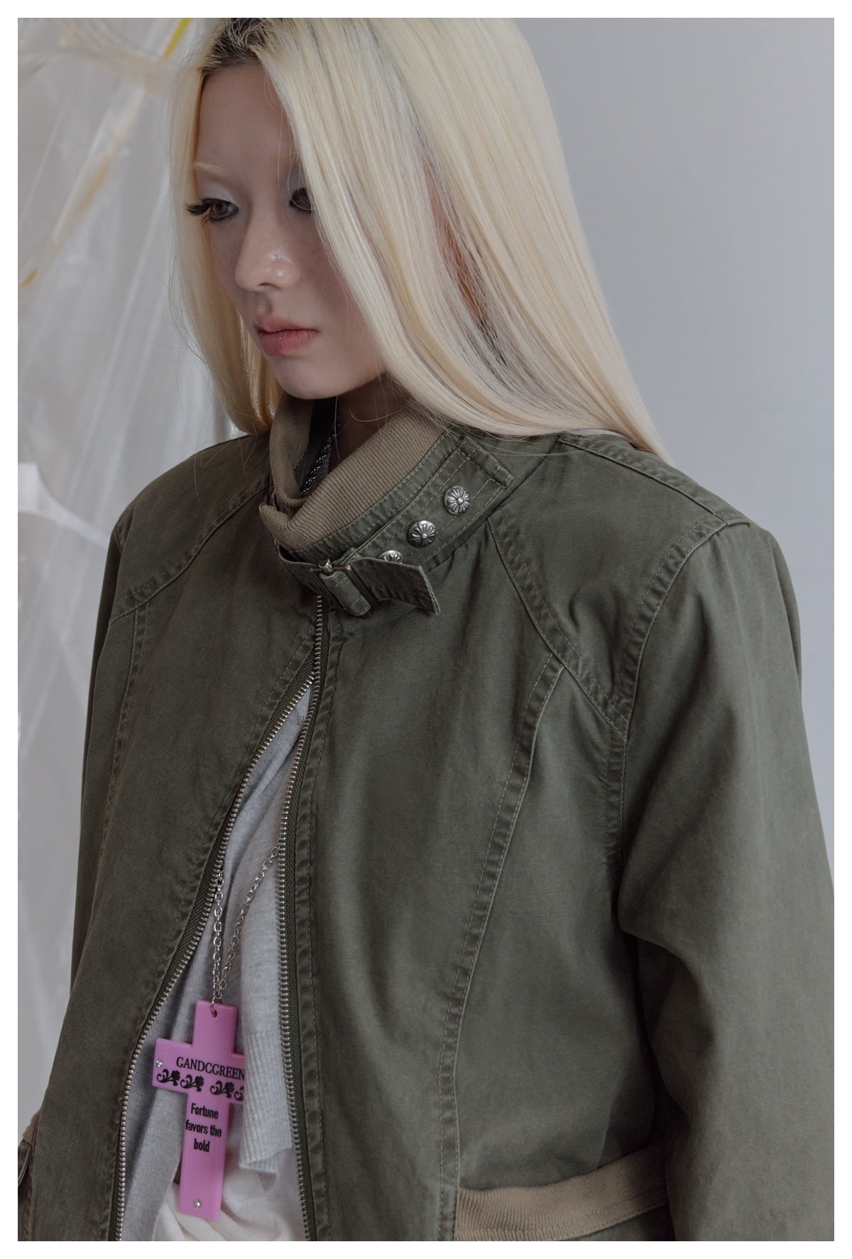 Close detail of an olive jacket's high collar, silver buttons, buckle, stitching and front zipper, worn by a model.