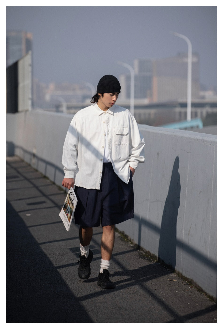 Model walking in a white button-up shirt and loose navy shorts, with a black beanie and sneakers.