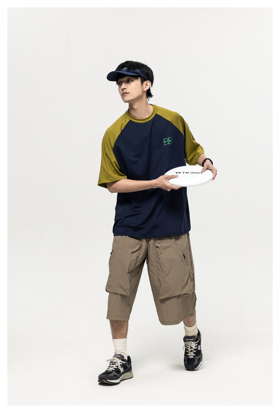 Model wearing a navy raglan T-shirt with olive sleeves and beige cargo shorts, holding a white flying disc.